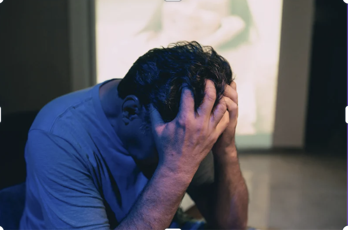 man in grey shirt sitting down with his head in his hands, in distress